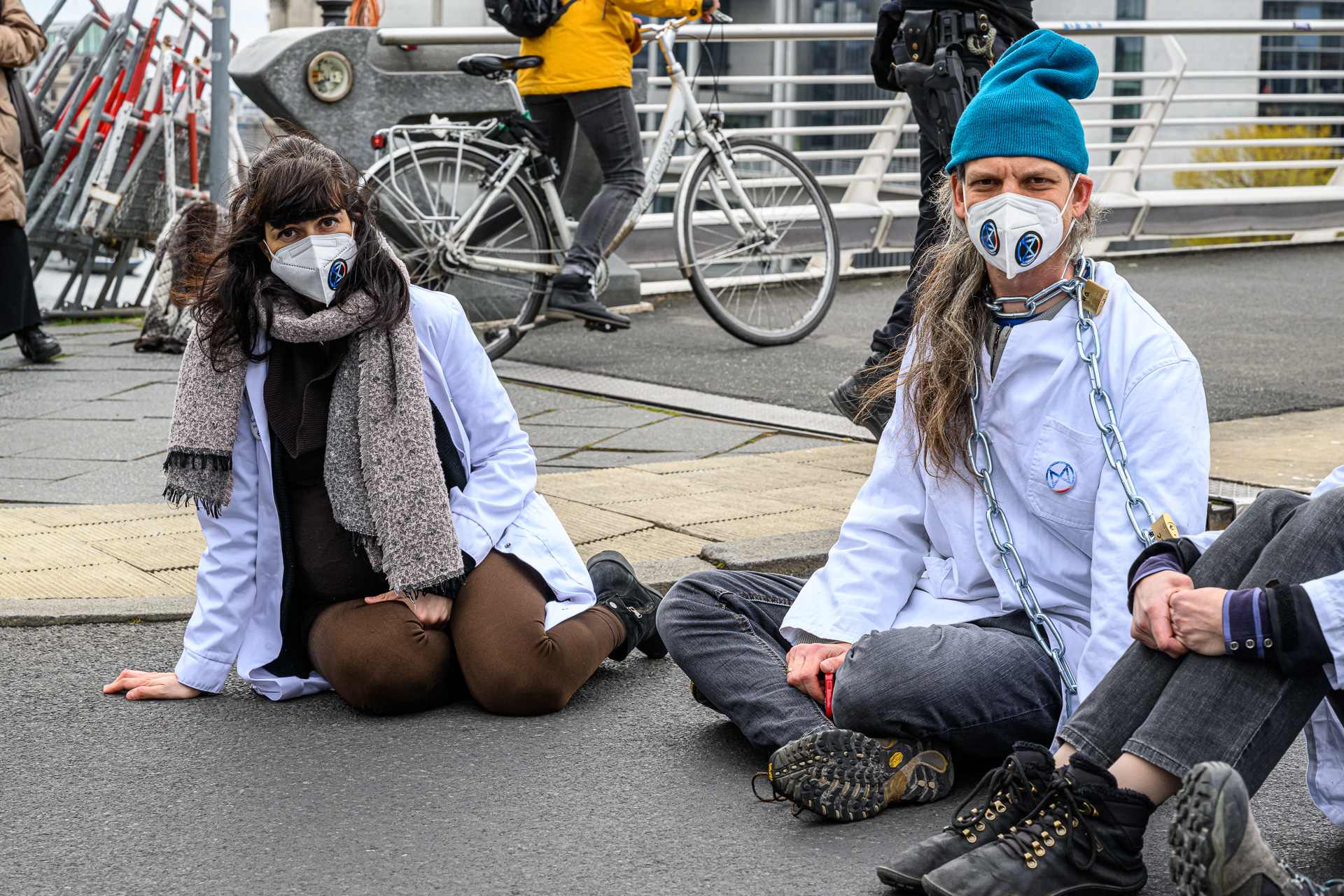 Menschen sitzen mit Laborkitteln auf der Straße im Regierungsviertel. Sie tragen Masken mit den Warming Stripes und dem Zeichen von Scientist Rebellion.