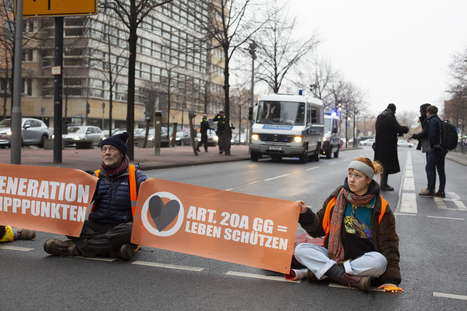 2 Menschen, Mio und Michael sitzen auf der Straße. Sie halten Banner, auf denen steht „Letzte Generation vor den Kipppunkten“ und „Artikel 20a Grundgesetz = Leben schützen“. Hinter ihnen sind Journalisten und Polizeiautos.
