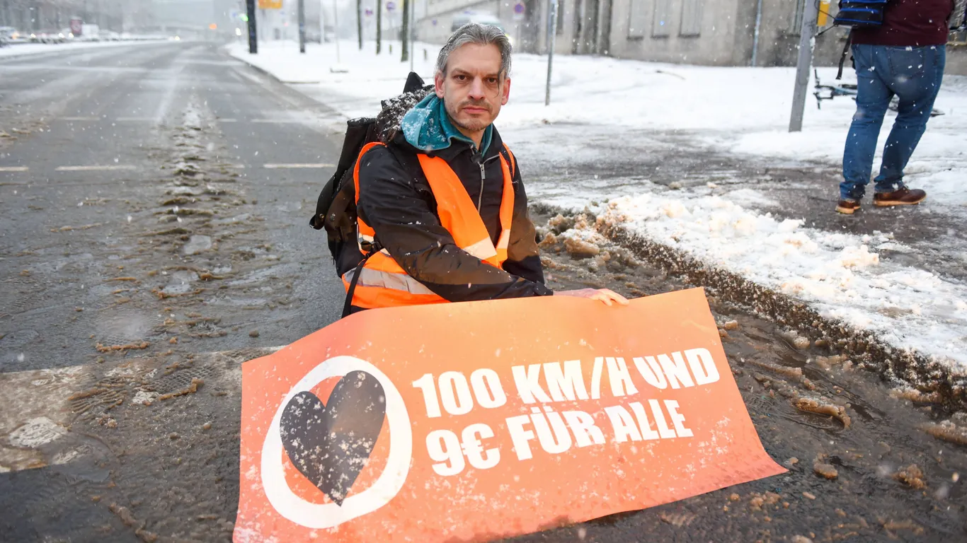 Christian Bläul sitzt auf einer Straße mit Schneematsch und hält ein Banner 100 km pro h und 9 € für alle; Foto: LausitzNews/imago-images-bilder
