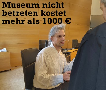 Christian Bläul sitzt im Landgericht. Über ihm ist Schrift zu sehen „Museum nicht betreten kostet mehr als 1000 €“