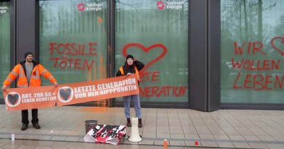 Arne und Karla stehen vor dem Hauptgebäude der SachsenEnergie. Sie halten Banner Artikel 20a Grundgesetz = Leben schützen und Letzte Generation vor den Kipppunkten. An den Scheiben sind Grafitti „Fossile töten“ und „Wir wollen leben”.